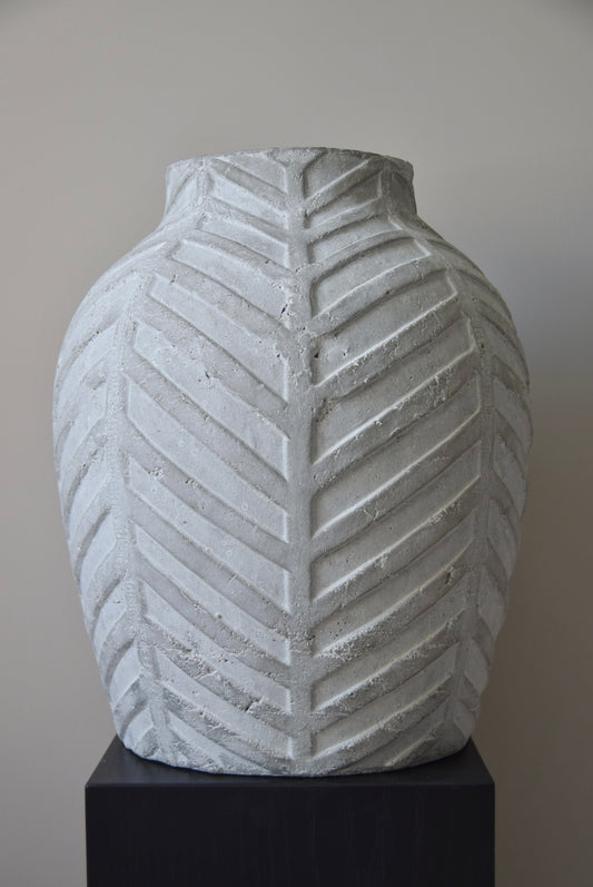 Grey chevron stone ceramic vase displayed on a black pedestal against a beige wall.