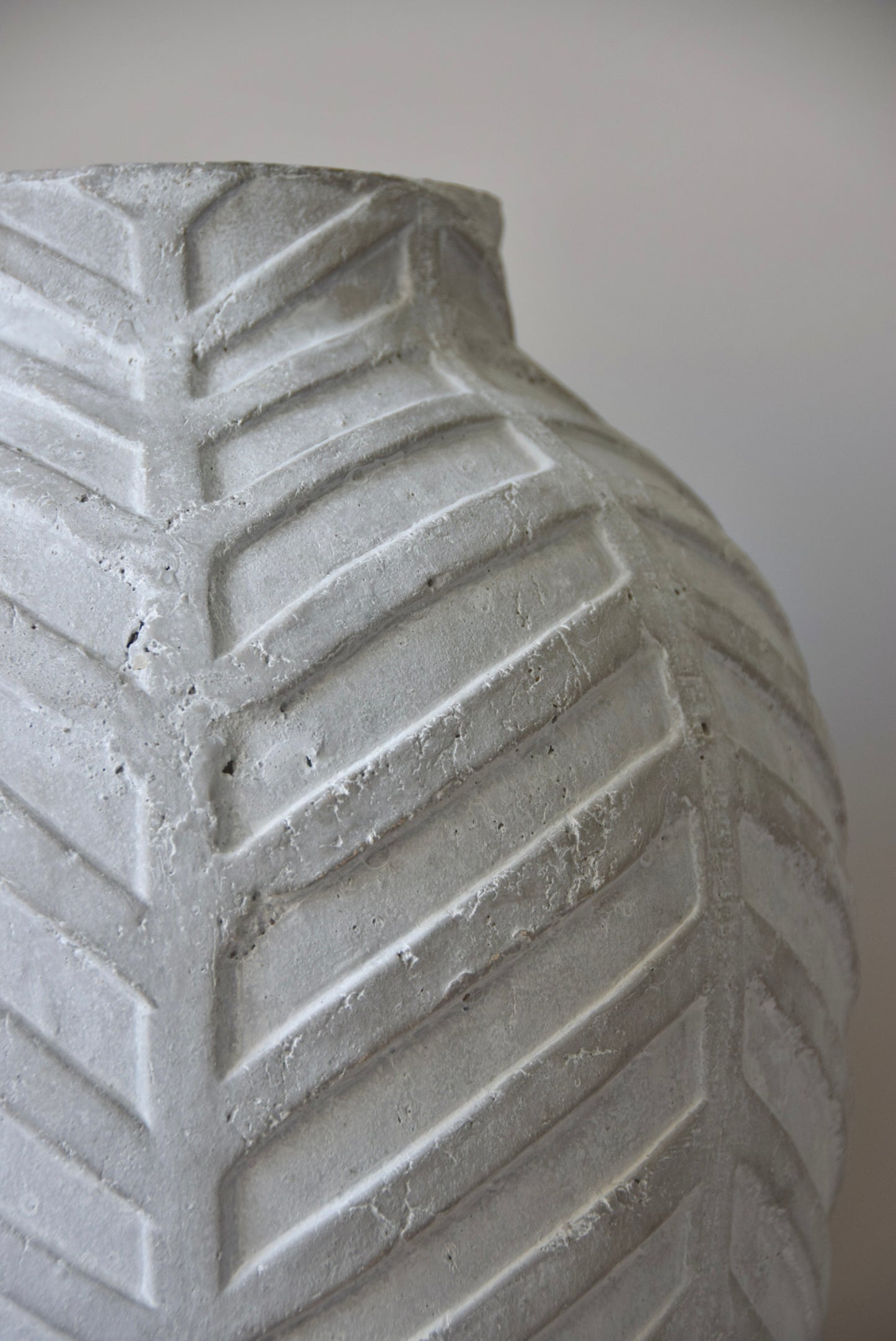 Grey chevron stone ceramic vase displayed on a black pedestal against a beige wall.