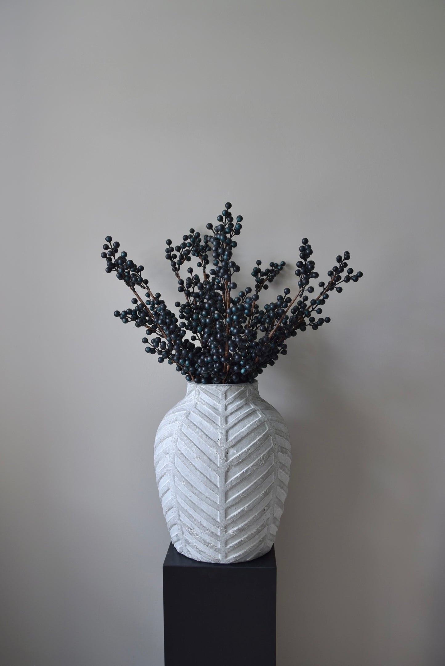 Stone chevron style vase placed on a black pedestal displaying some blueberry stems.