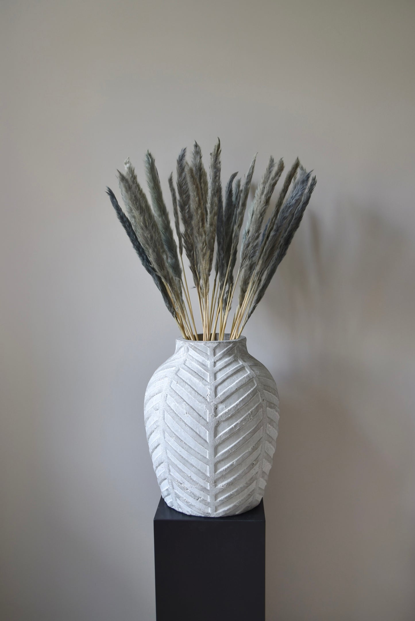 Grey reed pampas stems displayed in a chevron stone vase on a black pedestal.