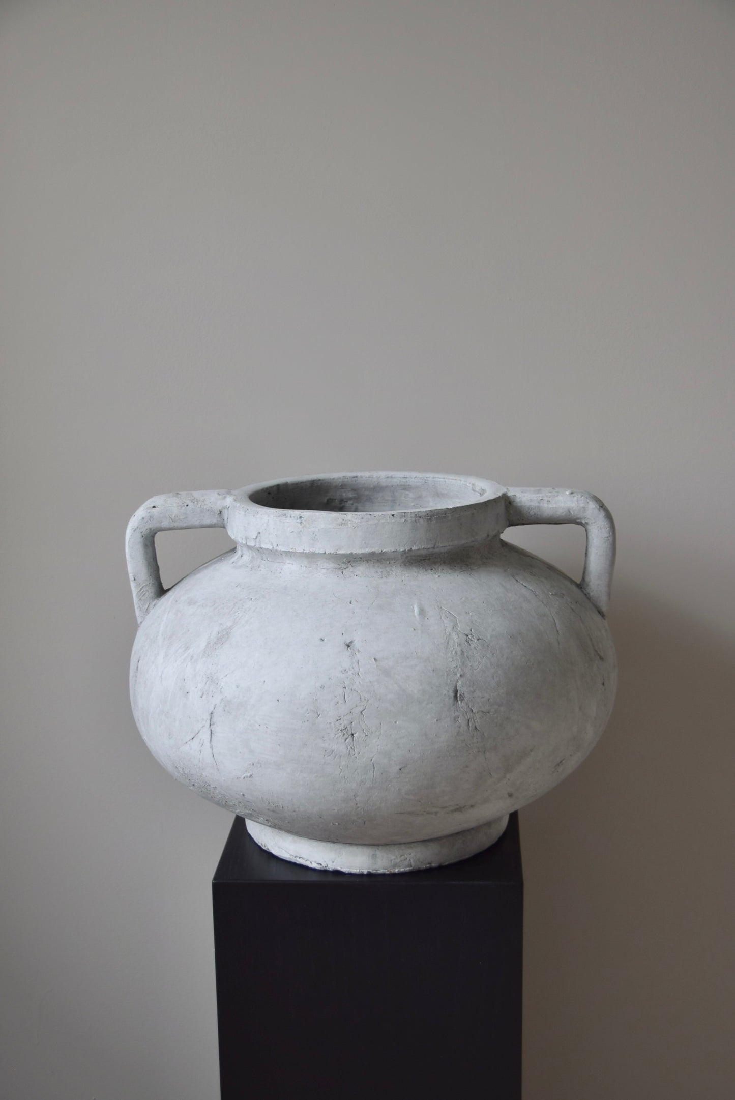 Grey stone Greek pelike vase with two handles displayed on a black pedestal.