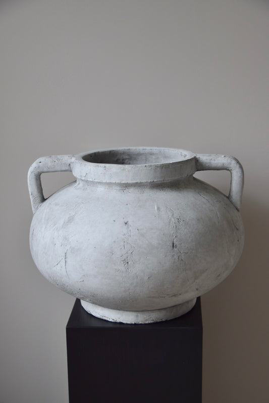 Grey stone Greek pelike vase with two handles displayed on a black pedestal.