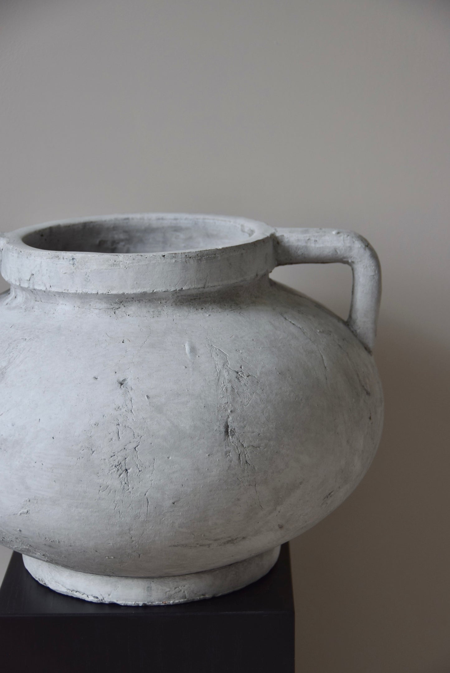 Grey stone Greek pelike vase with two handles displayed on a black pedestal.