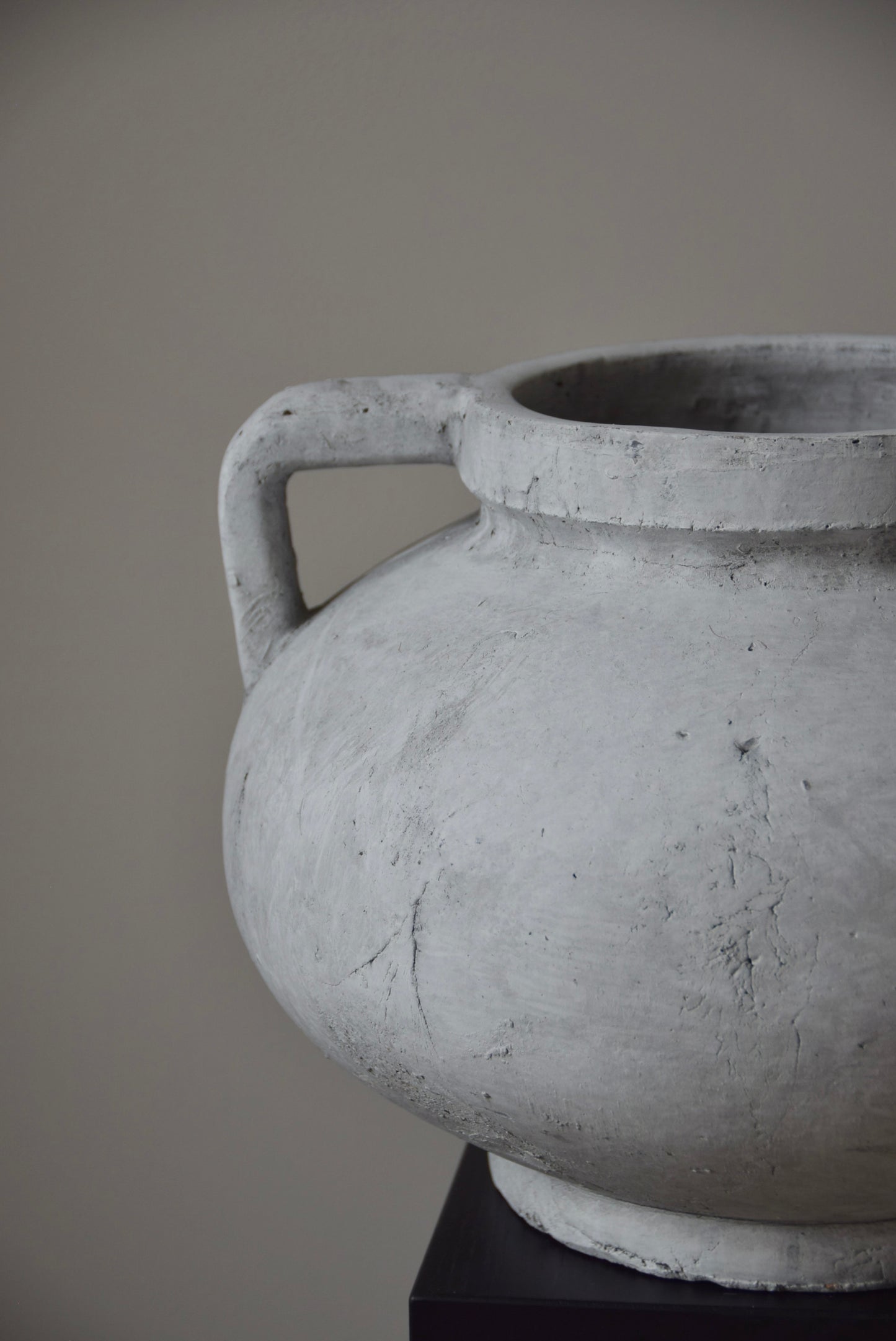 Grey stone Greek pelike vase with two handles displayed on a black pedestal.