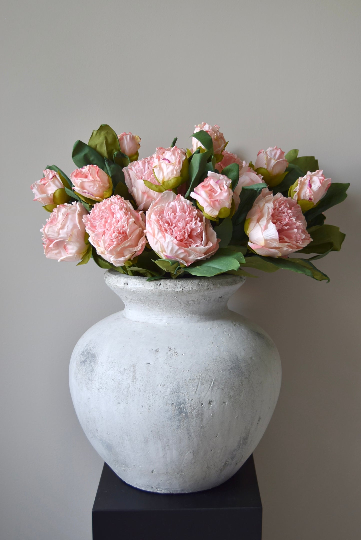 Stone vase placed on a black pedestal against a neutral coloured wall displaying a lovely bunch of pale pink peonies.