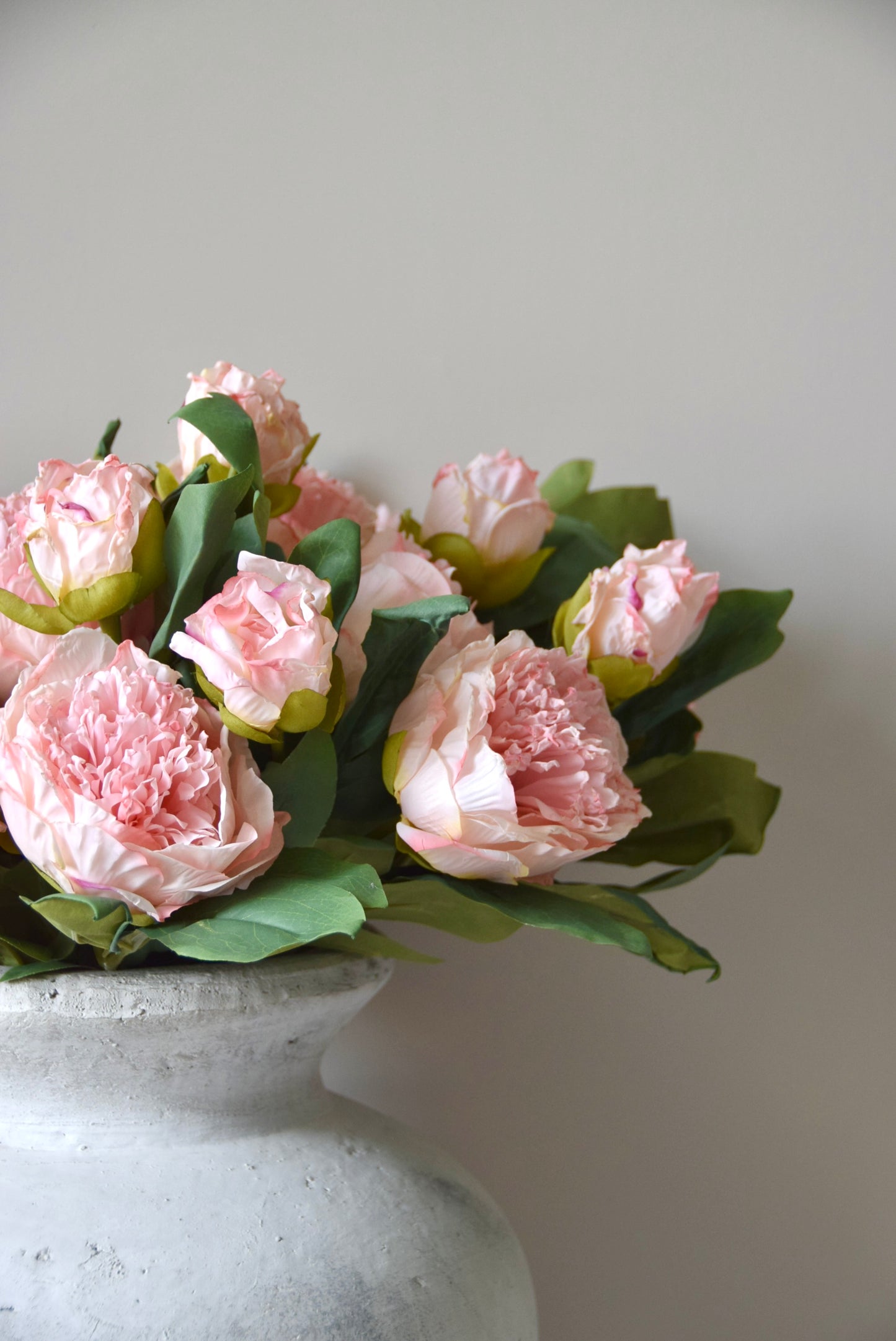Stone vase placed on a black pedestal against a neutral coloured wall displaying a lovely bunch of pale pink peonies.