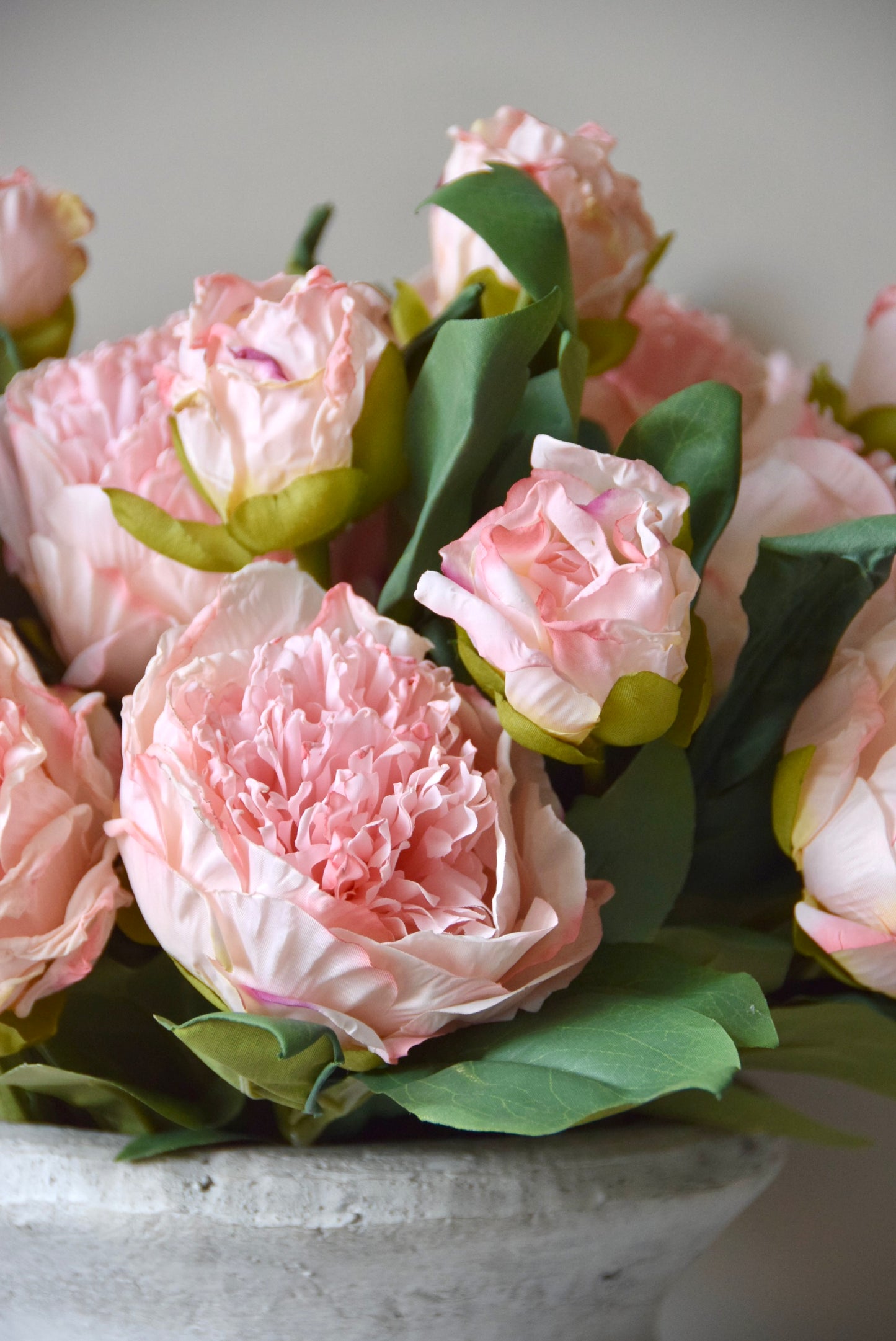 Stone vase placed on a black pedestal against a neutral coloured wall displaying a lovely bunch of pale pink peonies.