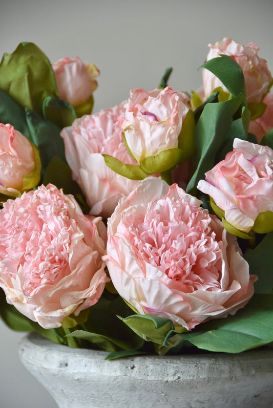 Stone vase placed on a black pedestal against a neutral coloured wall displaying a lovely bunch of pale pink peonies.