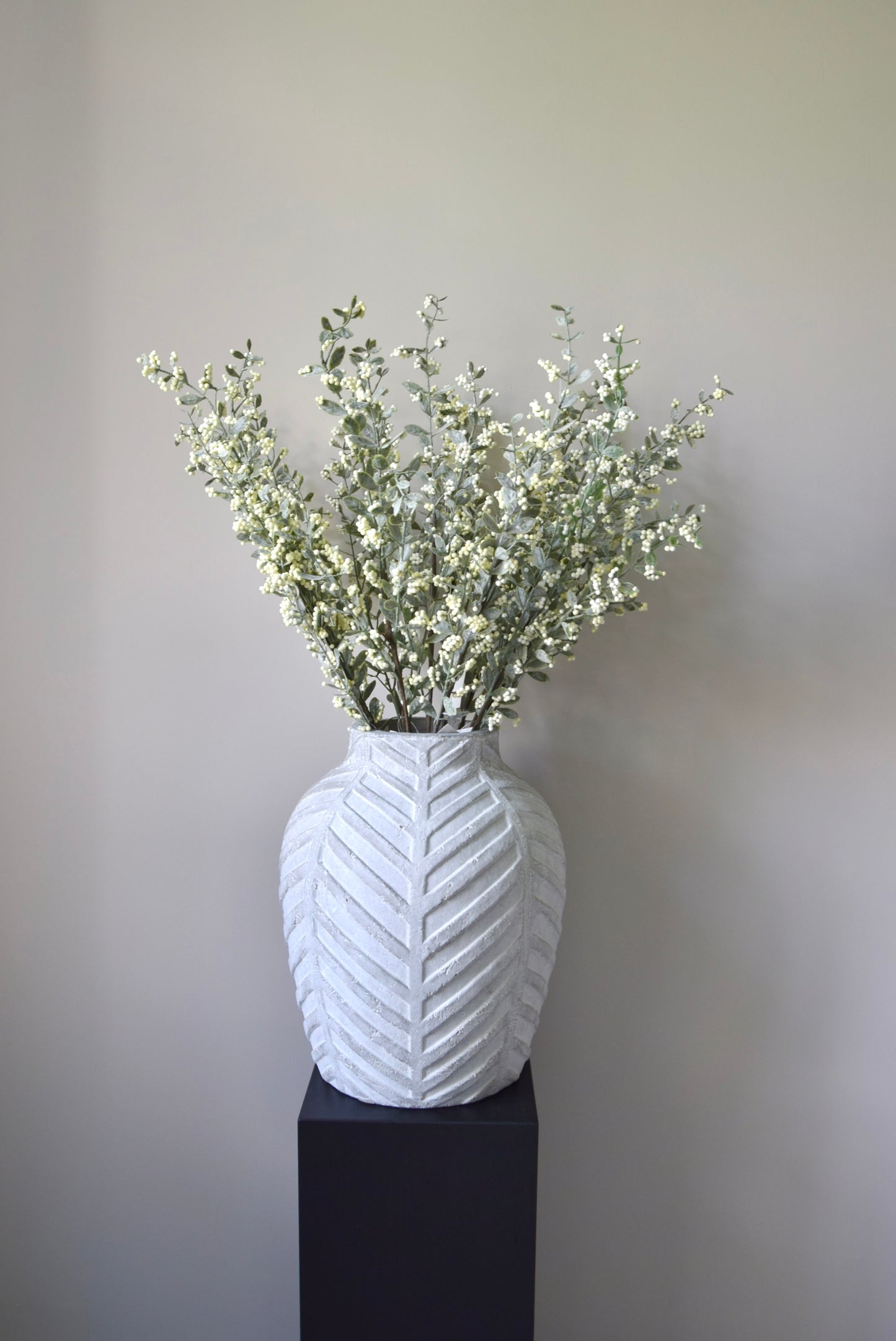 Stone chevron style vase placed on a black pedestal displaying a bunch of artificial berry leaf stems.