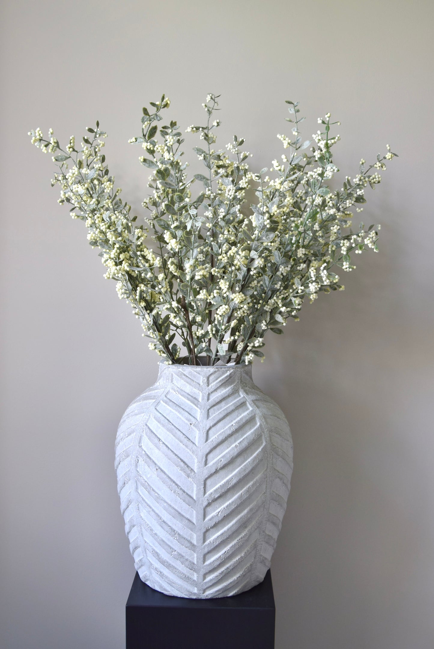 Stone chevron style vase placed on a black pedestal displaying a bunch of artificial berry leaf stems.