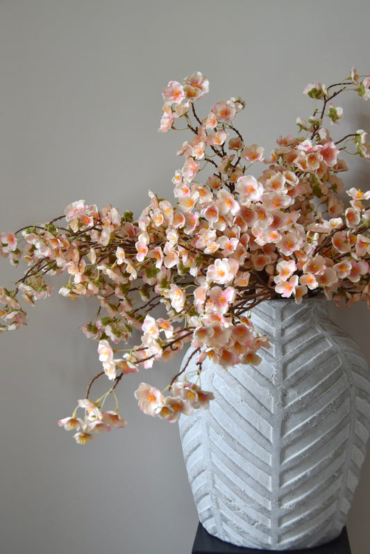 Stone chevron style vase placed on a black pedestal with a bunch of drooping pink plum blossom stems.