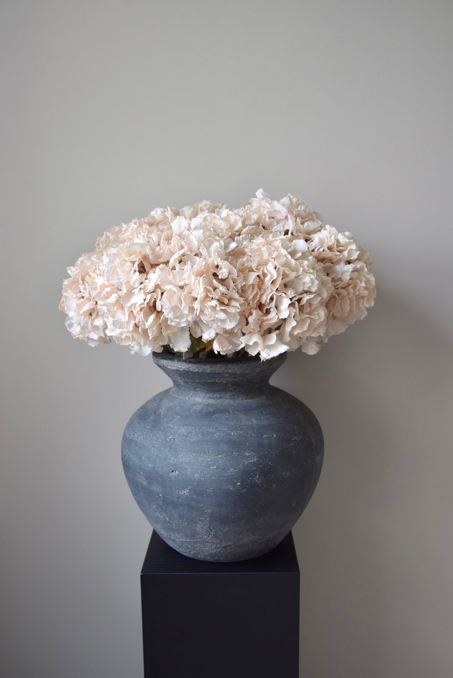 Grey vase with cream flowers on a black pedestal against a grey background