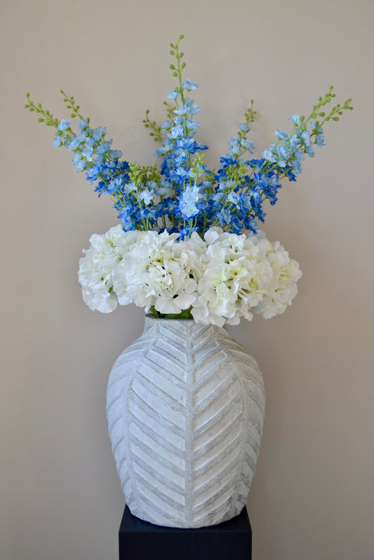 White artificial hydrangea and blue delphinium flower arrangement placed in a chevron style vase on a black pedestal against a plain wall.
