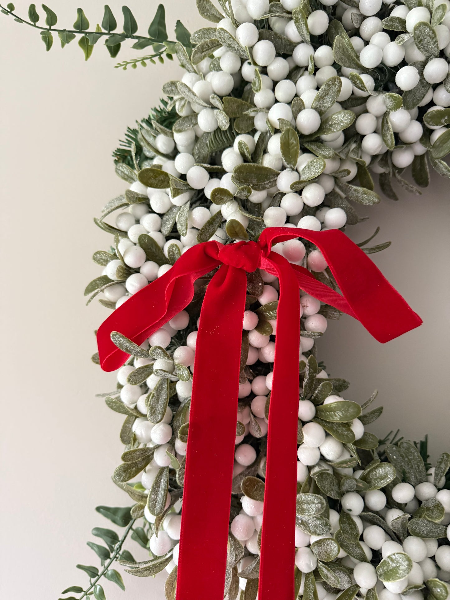 Decorative wreath with greenery, white berries, and a red ribbon on a neutral background