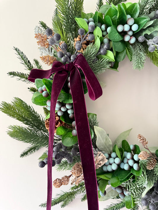 Decorative wreath with greenery, berries, and a burgundy ribbon on a light background