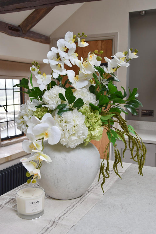 Stone antique vase placed on a kitchen countertop with an arrangement of white orchids, white hydrangeas, green hydrangeas and green foliage. Displayed in a country style kitchen for home decor.