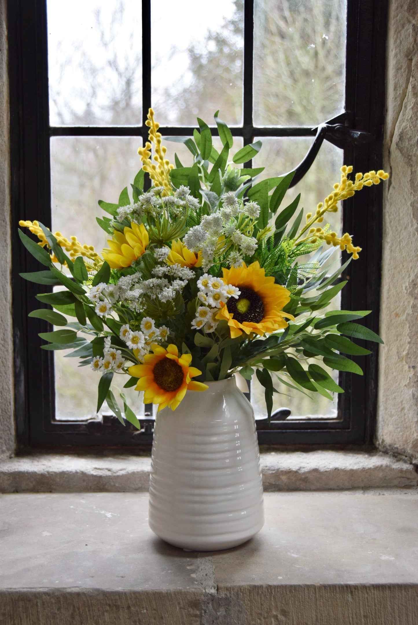 Cream country style vase placed on a stone windowsill with an arrangement of yellow sunflowers and green foliage. Displayed in a country style window for home decor.