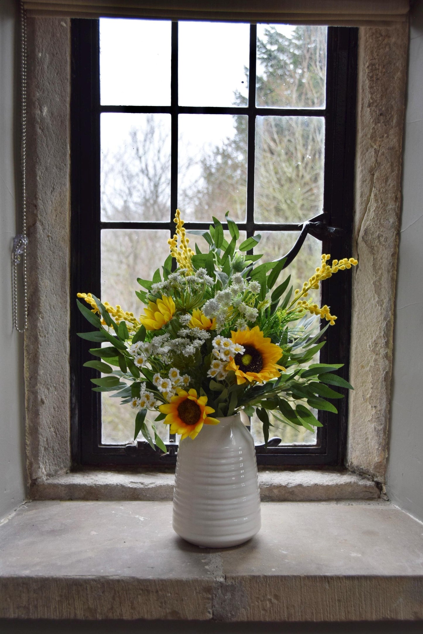 Cream country style vase placed on a stone windowsill with an arrangement of yellow sunflowers and green foliage. Displayed in a country style window for home decor.