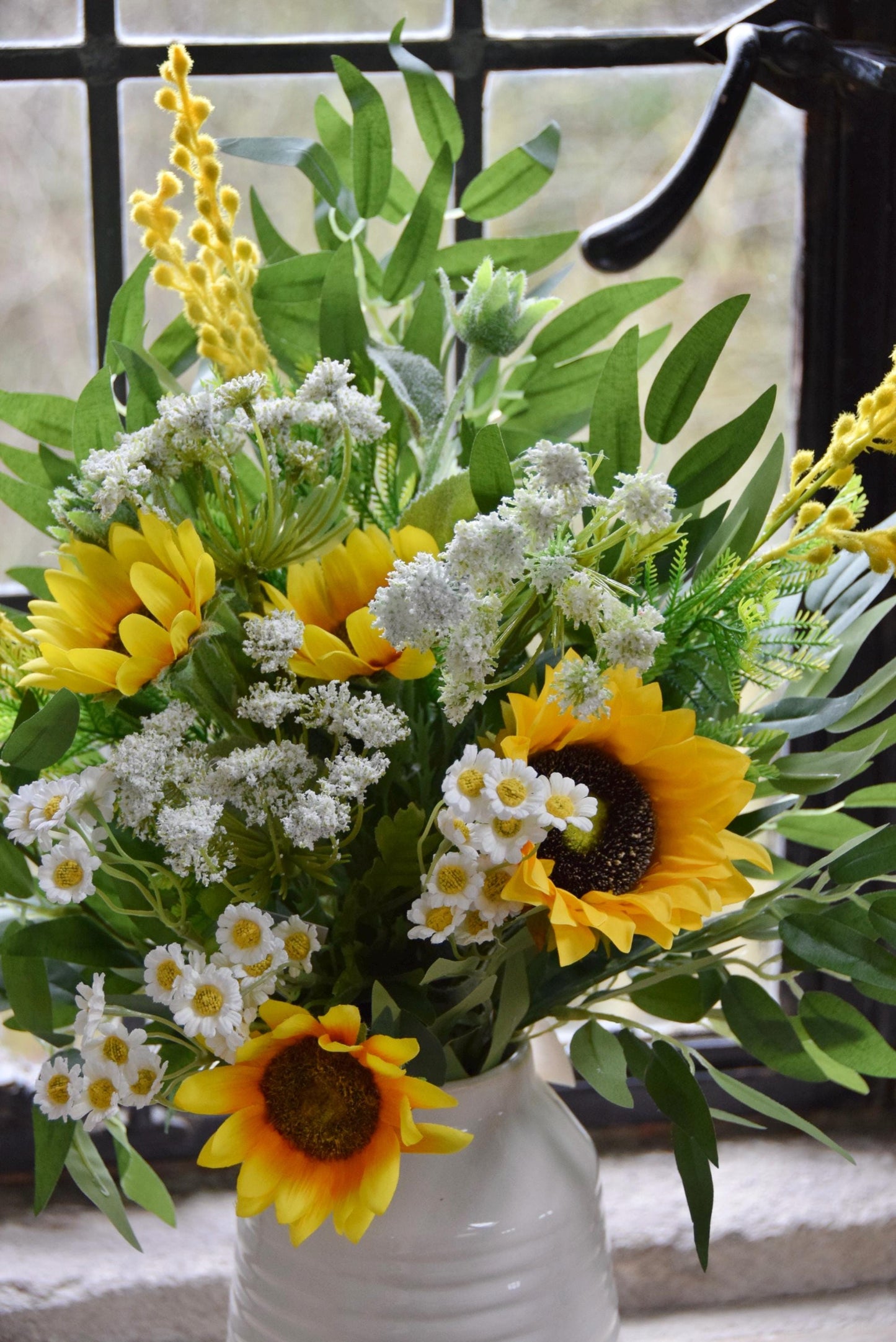 Cream country style vase placed on a stone windowsill with an arrangement of yellow sunflowers and green foliage. Displayed in a country style window for home decor.