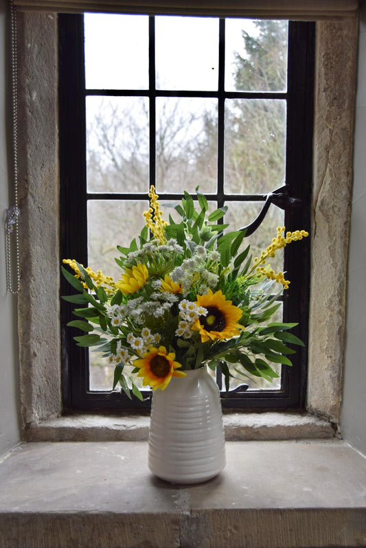 Cream country style vase placed on a stone windowsill with an arrangement of yellow sunflowers and green foliage. Displayed in a country style window for home decor.