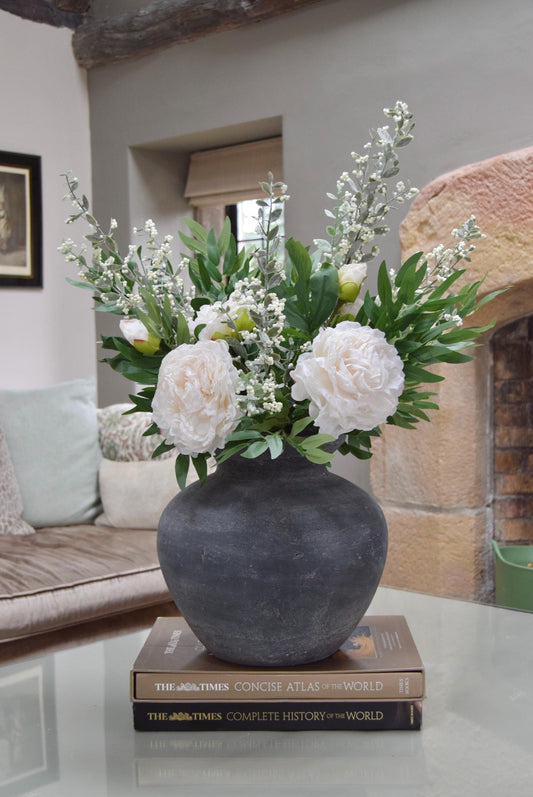 Grey antique vase placed on a coffee table with artificial ruscus foliage and faux cream peony flowers. Arranged in a country style living room for home decor.