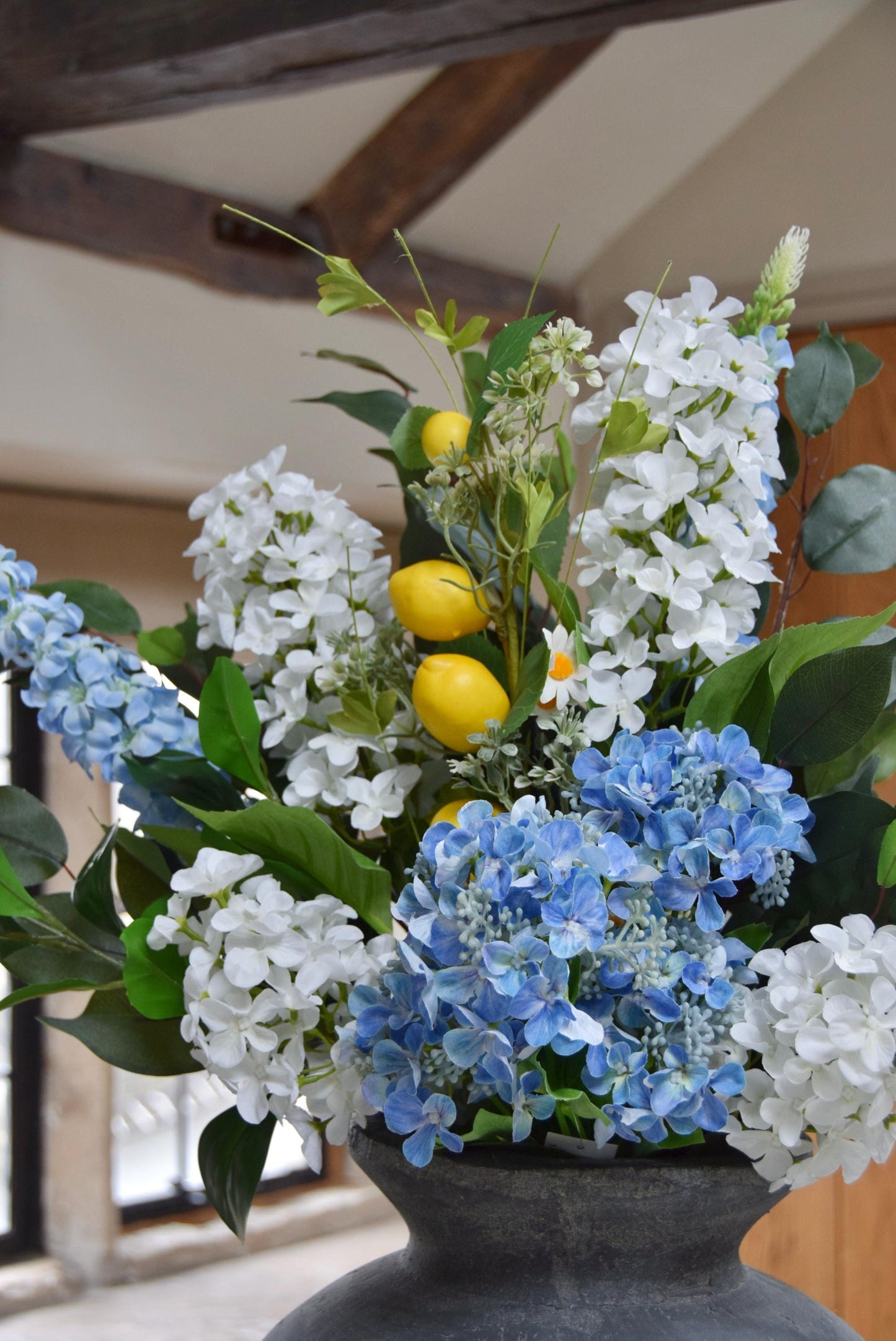 Grey antique vase placed on a kitchen countertop with arrangement of blue and white hydrangeas with lemons and green foliage. Displayed in a country style kitchen for home decor.