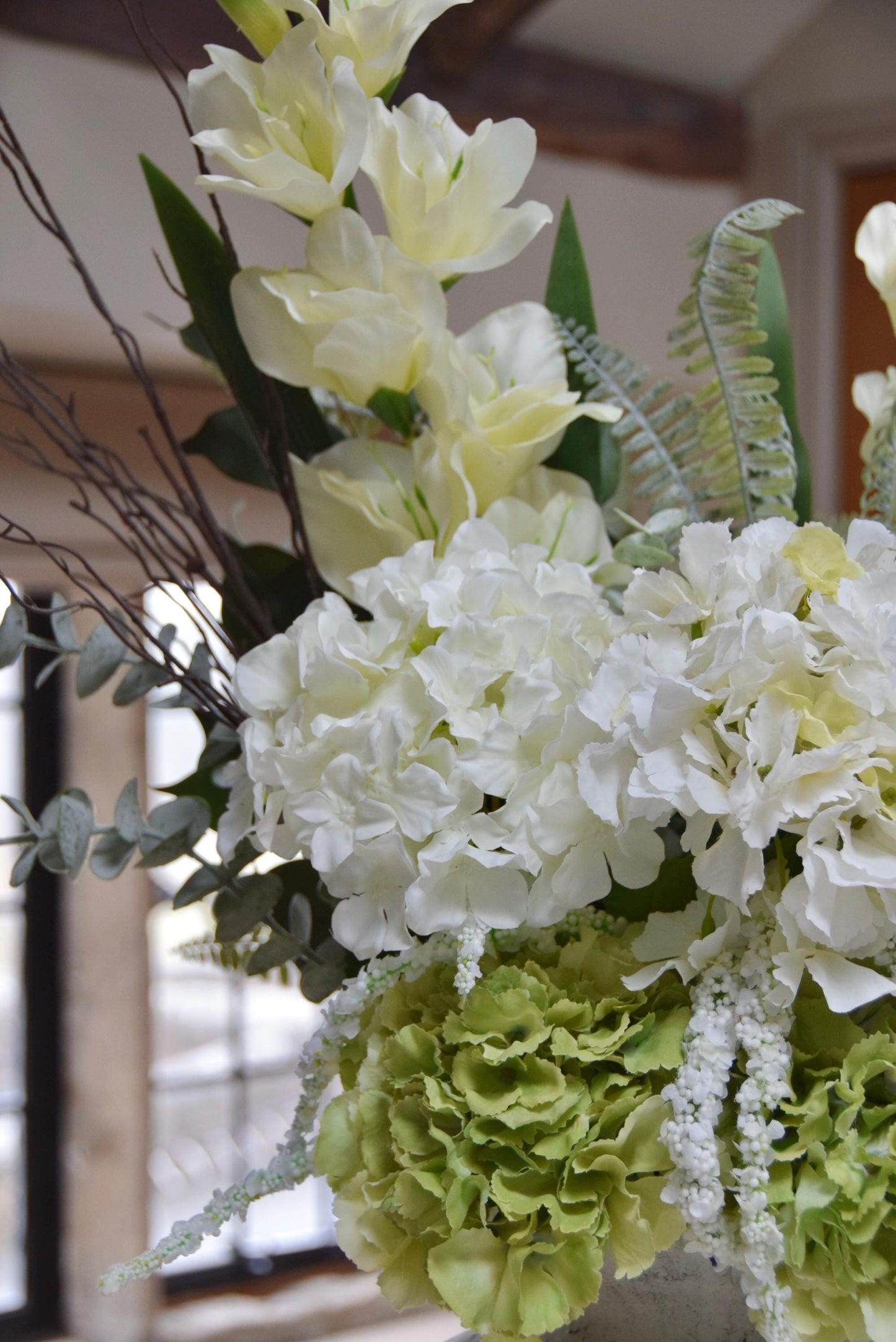 Stone antique vase placed on a kitchen countertop with an arrangement of white hydrangeas, green hydrangeas, cream gladioli and foliage. Displayed in a country style kitchen for home decor.