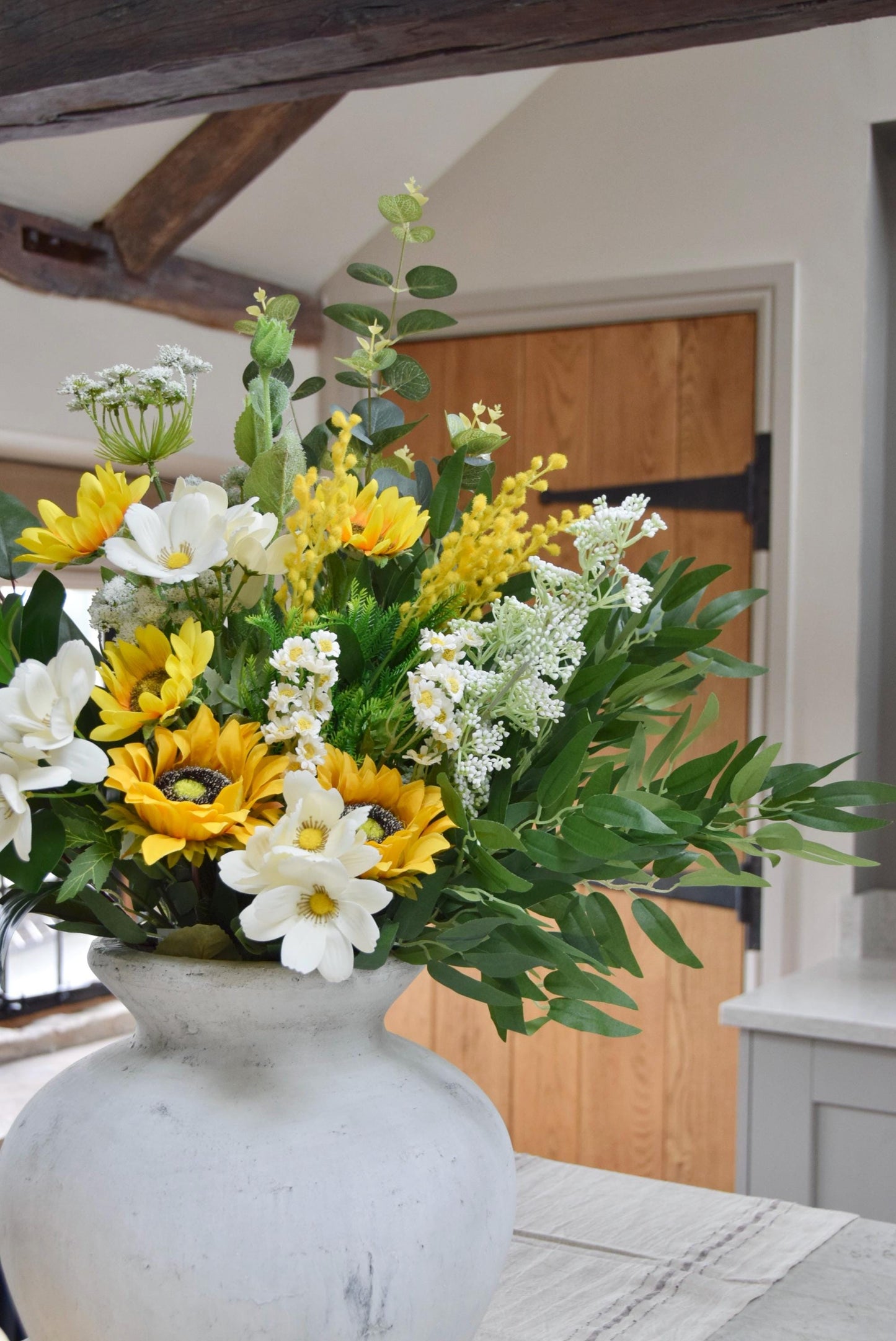 Stone antique vase placed on a kitchen countertop with an arrangement of yellow sunflowers, white wildflowers and green foliage. Displayed in a country style kitchen for home decor.