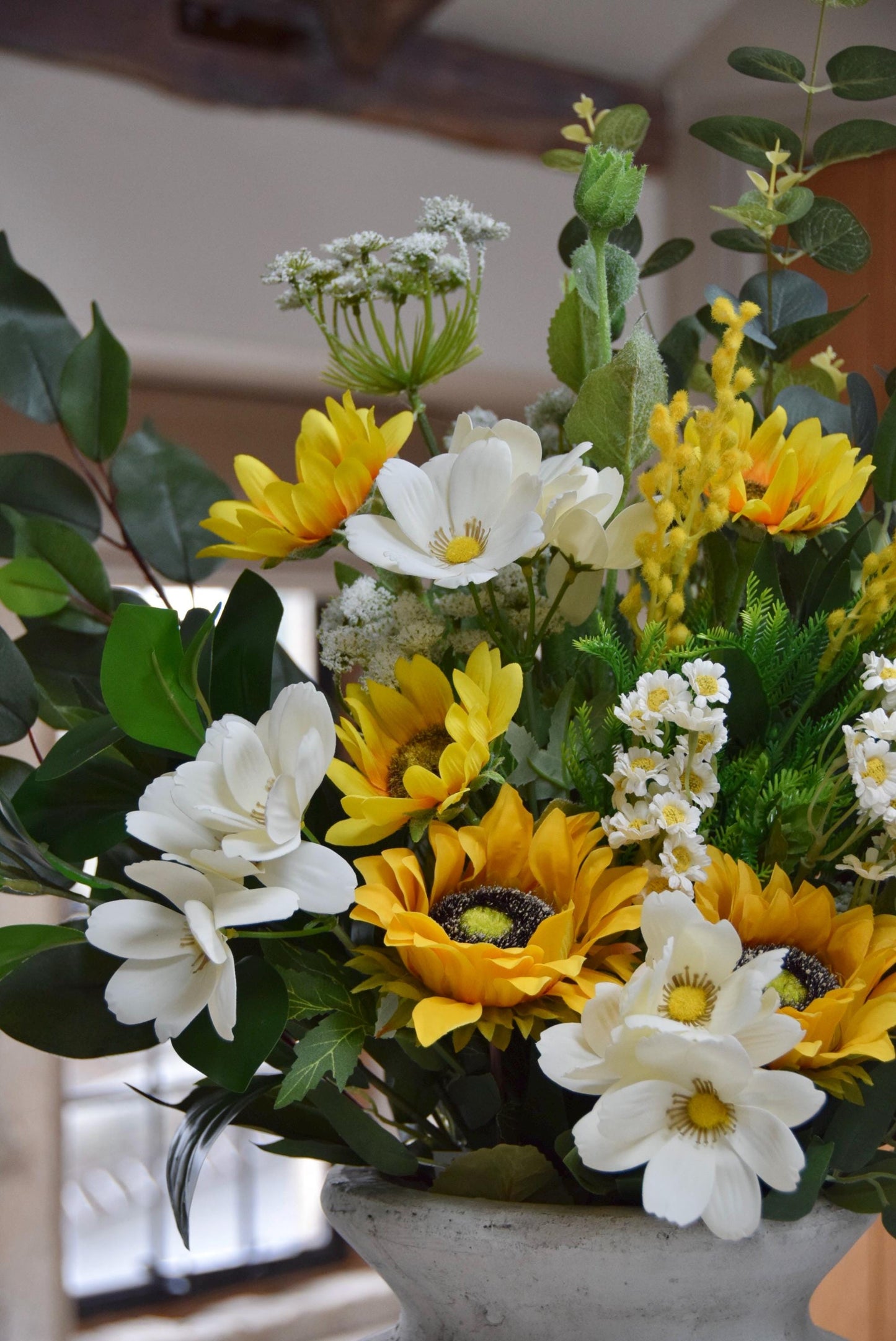 Stone antique vase placed on a kitchen countertop with an arrangement of yellow sunflowers, white wildflowers and green foliage. Displayed in a country style kitchen for home decor.