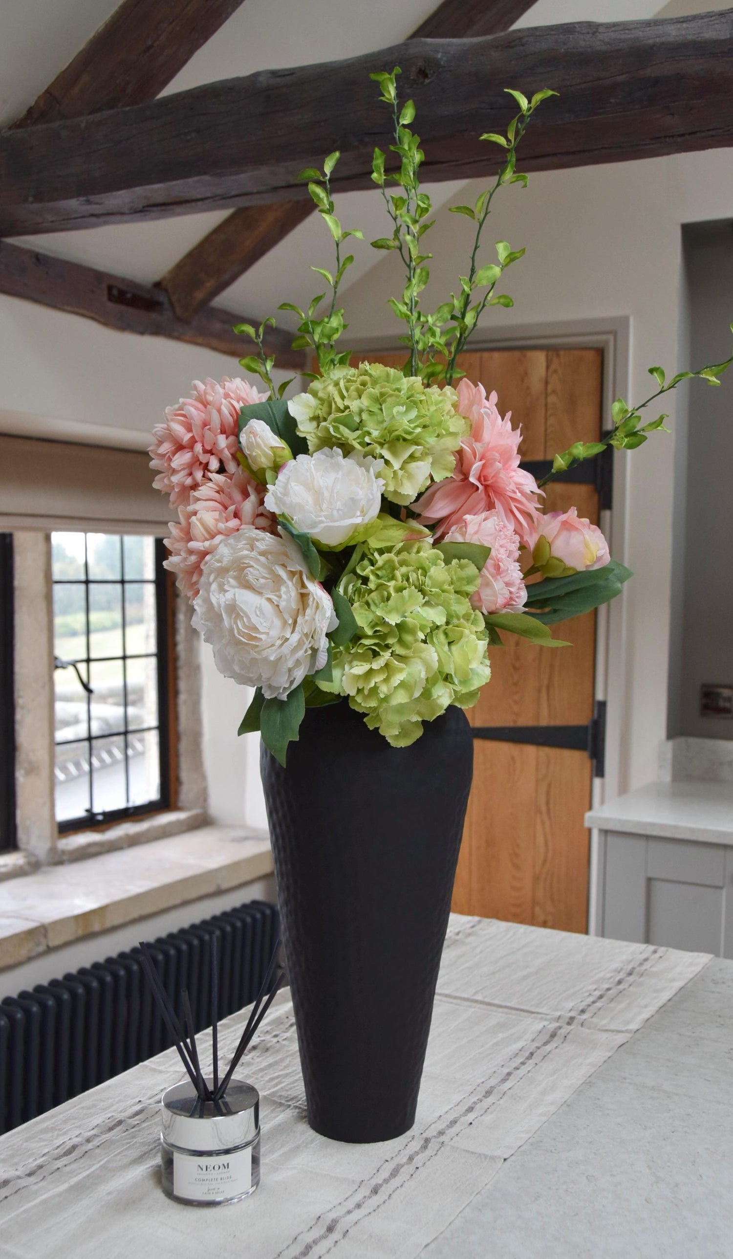 Tall black vase placed on a kitchen countertop with an arrangement of pink and green flowers including green hydrangeas, pink mum flowers, pink dahlias and white peonies with foliage. Displayed in a country style kitchen for home decor.