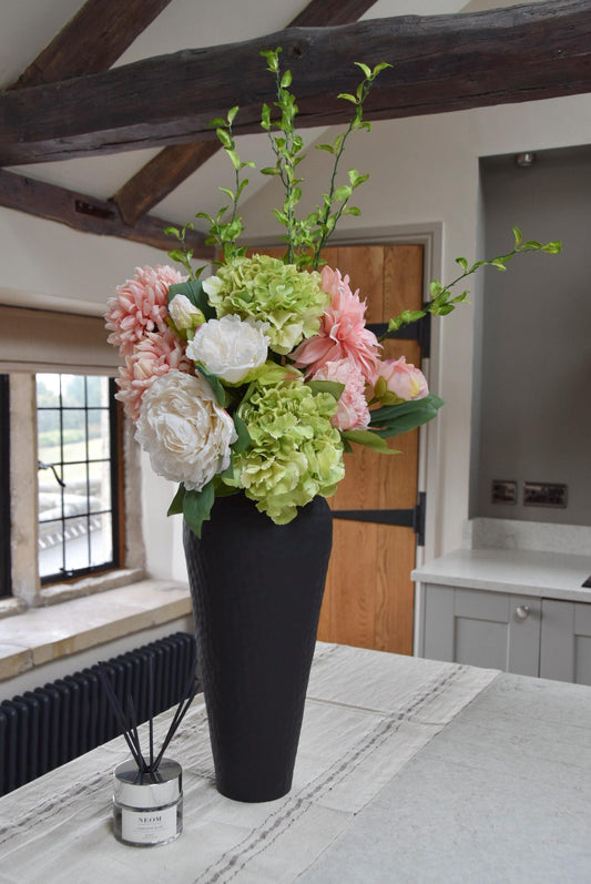 Tall black vase placed on a kitchen countertop with an arrangement of pink and green flowers including green hydrangeas, pink mum flowers, pink dahlias and white peonies with foliage. Displayed in a country style kitchen for home decor.