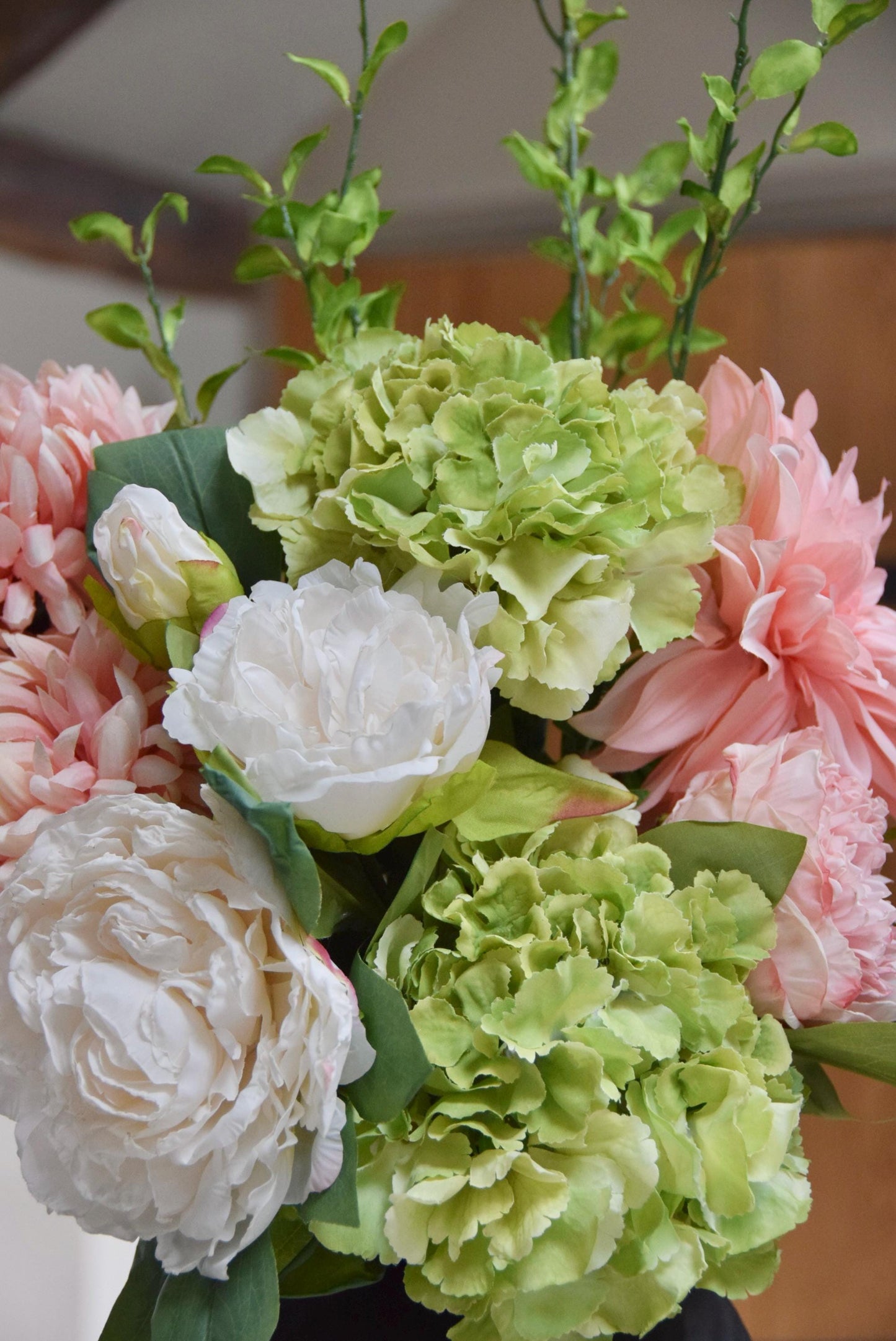 Tall black vase placed on a kitchen countertop with an arrangement of pink and green flowers including green hydrangeas, pink mum flowers, pink dahlias and white peonies with foliage. Displayed in a country style kitchen for home decor.