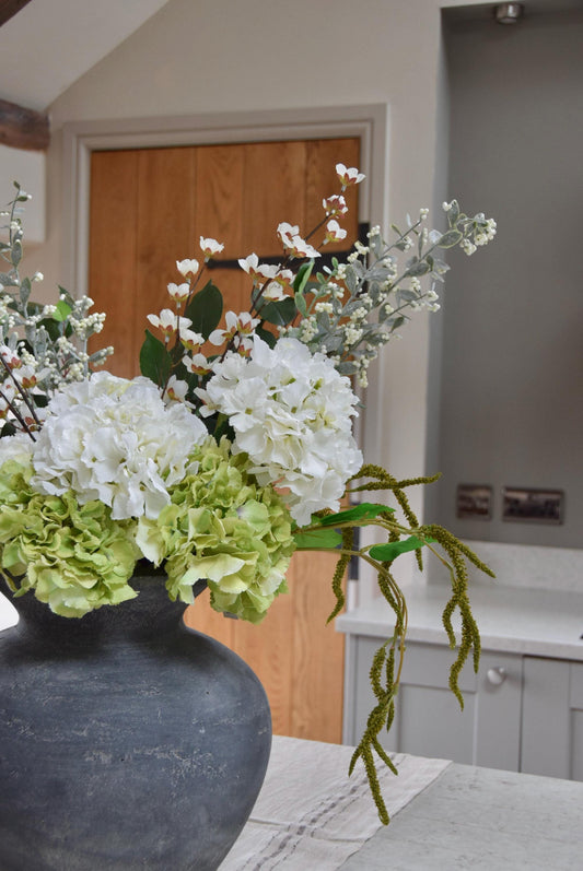 Grey vase placed on a kitchen countertop with a mixture of green and white hydrangeas with lots of mixed foliage. Displayed in a country style kitchen for home decor.
