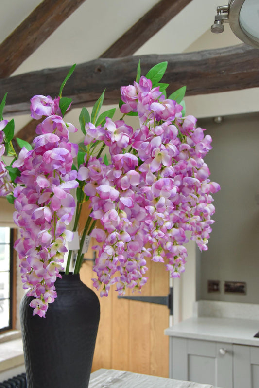 Tall black vase set on a kitchen countertop with a bunch of purple artificial wisteria stems. Displayed in a country style kitchen for home decor.