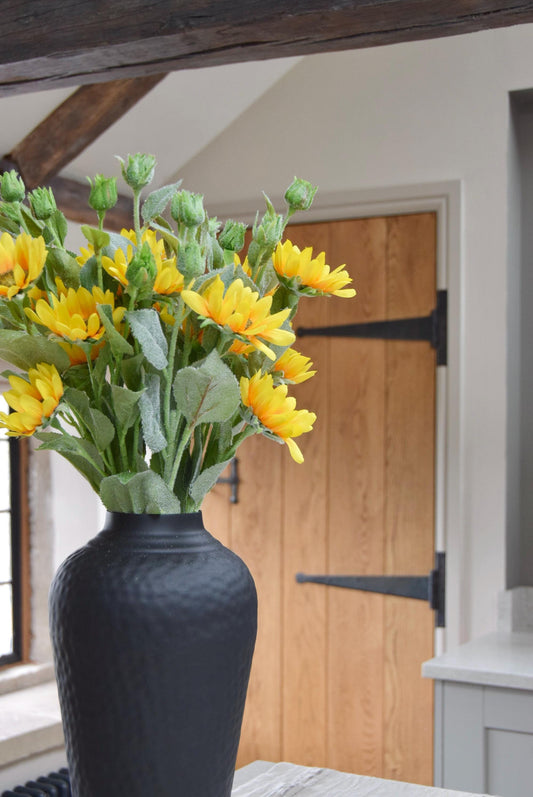 Tall black vase placed on a kitchen countertop with a bunch of artificial yellow sunflower sprays. Displayed in a country style kitchen for home decor.