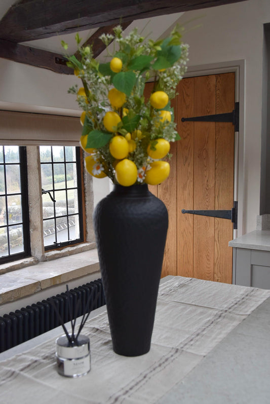 Tall black vase placed on a kitchen countertop with a bunch of faux lemon branches. Displayed in a country style kitchen for home decor.