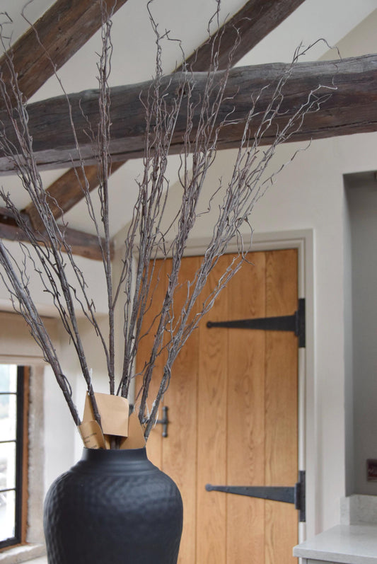 Tall black vase placed on a kitchen countertop with a bunch of faux brown branches. Displayed in a country style kitchen for home decor.