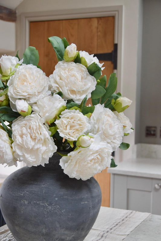 Grey antique vase placed on a kitchen countertop with a bunch of artificial cream peonies. Displayed in a country style kitchen for home decor.