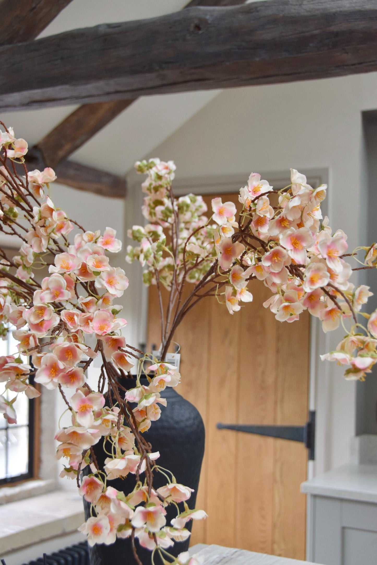 Tall black vase placed on a kitchen countertop with a bunch of hanging pink plum blossom stems. Displayed in a country style kitchen for home decor.