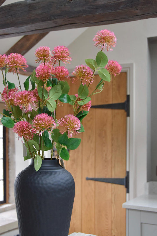 Tall black vase placed on a kitchen countertop with a bunch of pink pom flower stems. Displayed in a country style kitchen for home decor.