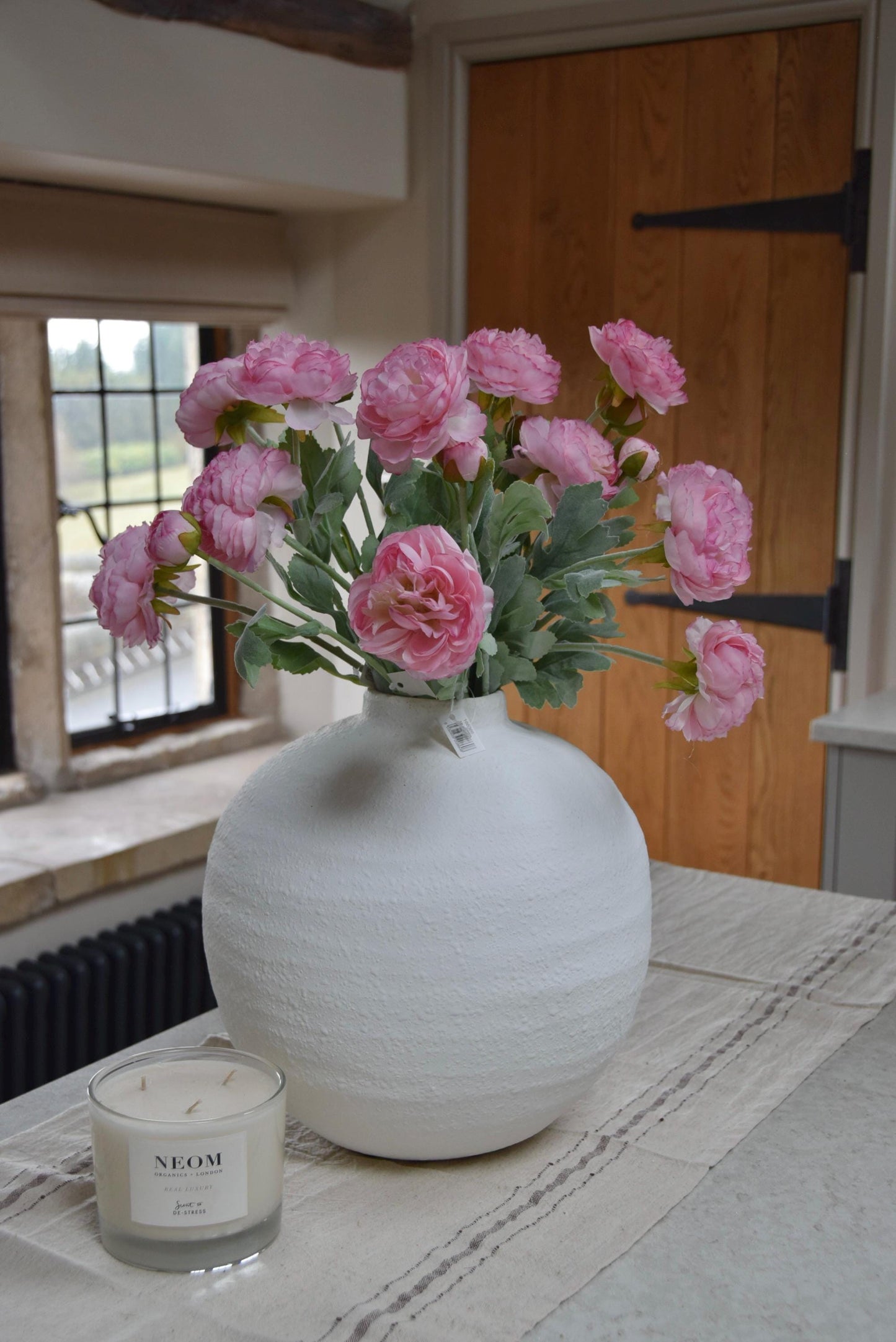White round vase placed on a kitchen countertop with a bunch of pink ranunculus stems. Displayed in a country style kitchen for home decor.
