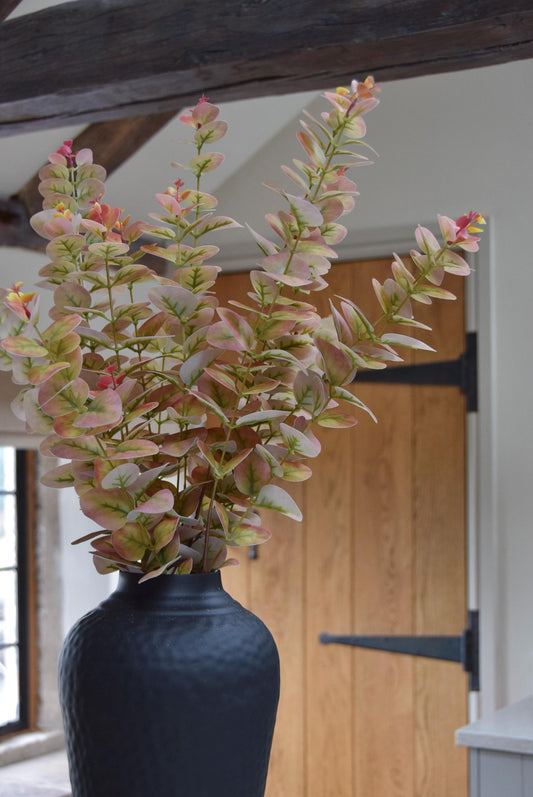 Tall black vase placed on a kitchen countertop with a bunch of pink and green blush eucalyptus stems. Displayed in a country style kitchen for home decor.