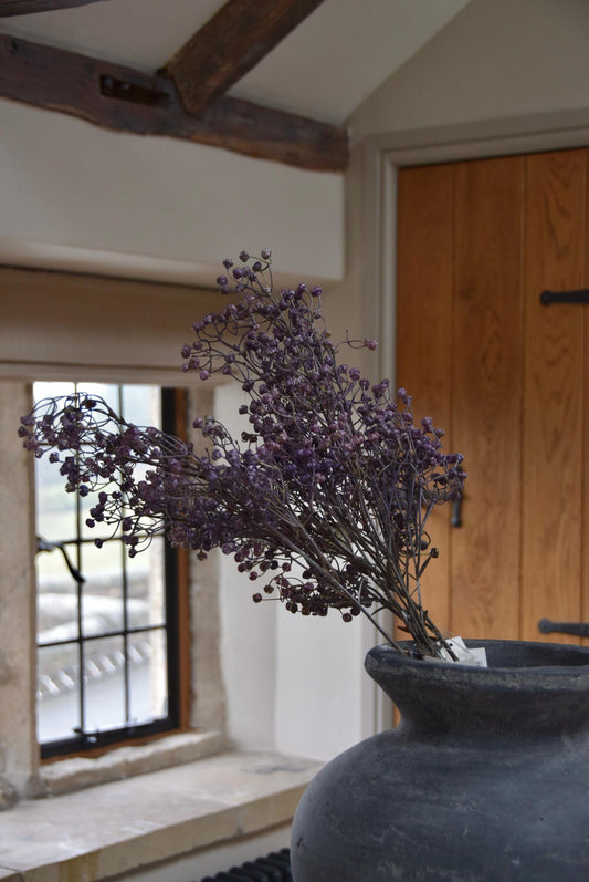 Grey antique vase with a bunch of purple dried look linum sprays placed on a kitchen countertop. Displayed in a country style kitchen for home decor.