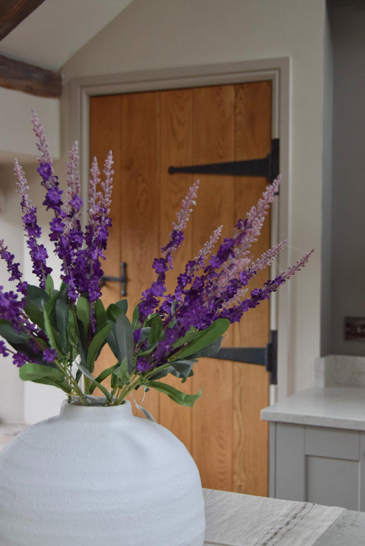 White round vase with a bunch of purple wild flower stems placed on a kitchen countertop. Displayed in a country style kitchen for home decor.