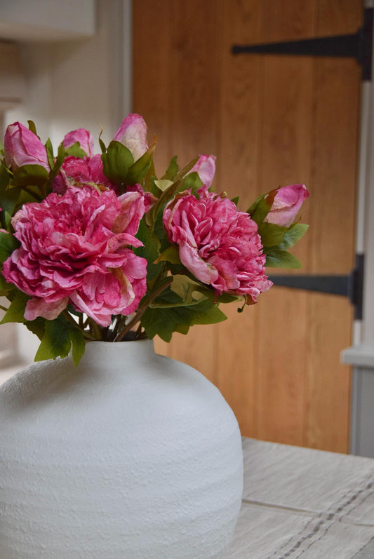White round vase placed on a kitchen countertop with a bunch of hot pink peonies. Displayed in a country style kitchen for home decor.