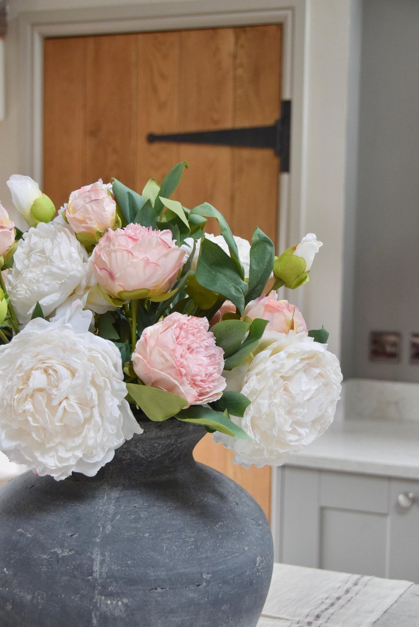Grey antique vase placed on a kitchen counter top with an arrangement of pink and cream peonies. Arranged in a country style kitchen for home decor.