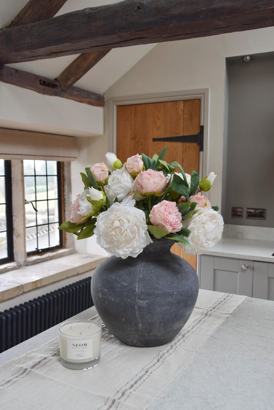 Grey antique vase placed on a kitchen counter top with an arrangement of pink and cream peonies. Arranged in a country style kitchen for home decor.