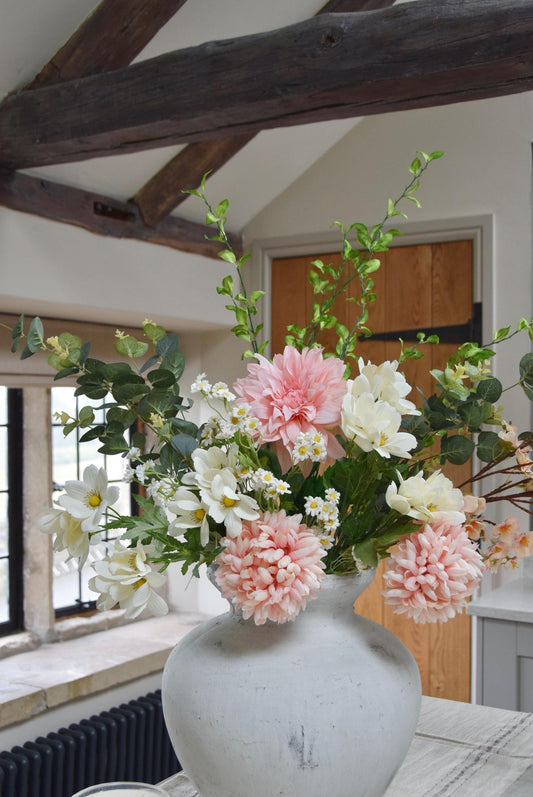 Stone antique vase placed on a kitchen counter top with an arrangement of pink dahlias and mum flowers with white wildflowers and green foliage. Set in a country style kitchen for home decor.