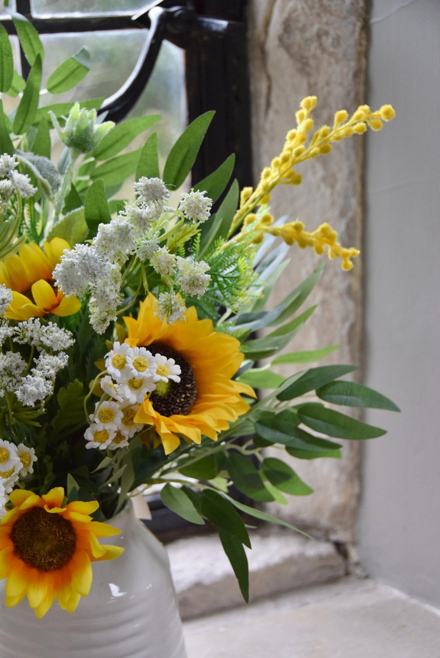 Cream country style vase placed on a stone windowsill with an arrangement of yellow sunflowers and green foliage. Displayed in a country style window for home decor.