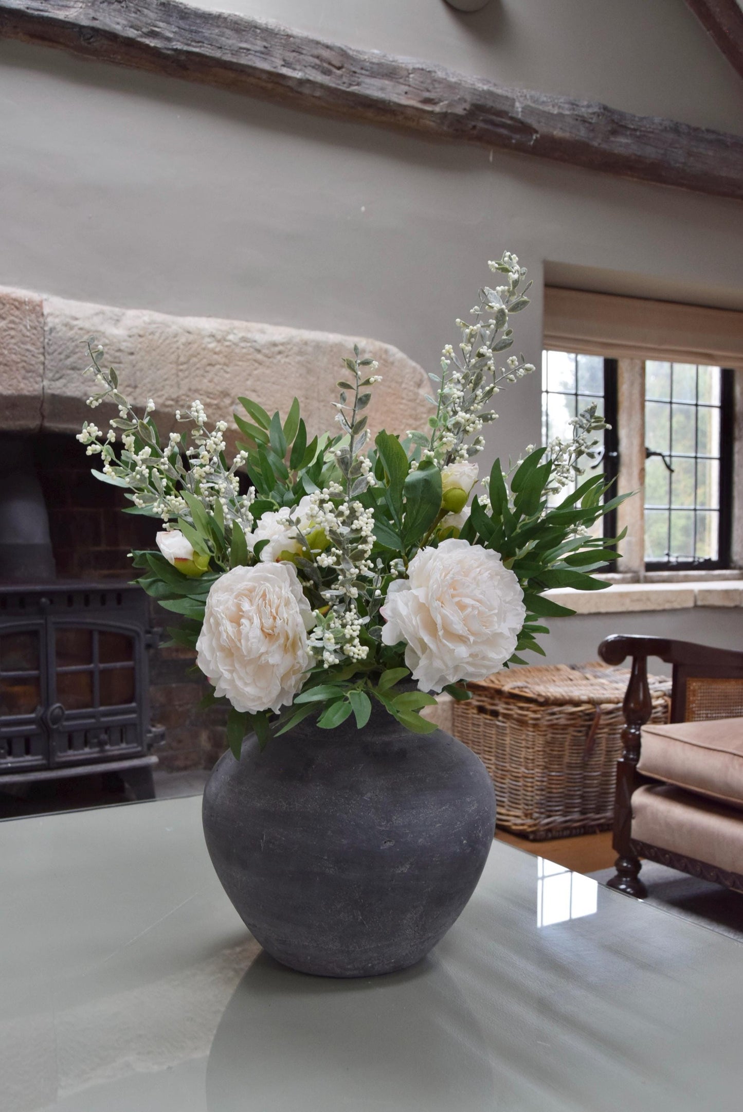 Grey antique vase placed on a coffee table with artificial ruscus foliage and faux cream peony flowers. Arranged in a country style living room for home decor.